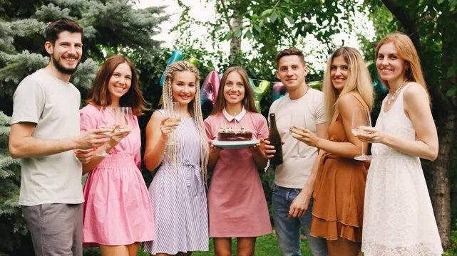 Woman with birthday cake with lit candles stands surrounded by her friends in front of festive decorations