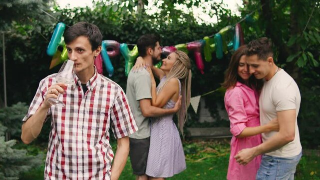 A lonely guy standing at the edge of a birthday party, sipping champagne while couples dance happily around him, highlighting his solitude amidst the celebration