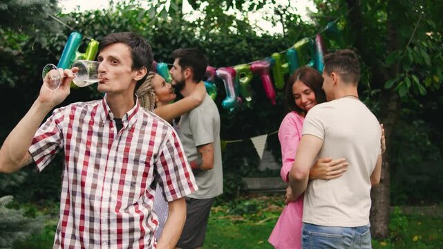 A lonely guy standing at the edge of a birthday party, sipping champagne while couples dance happily around him, highlighting his solitude amidst the celebration