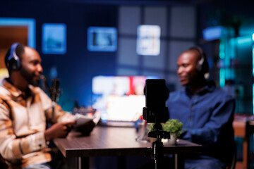 The podcast host uses smartphone on a tripod to record guests giving fans fascinating stories from living room. Invited to the home studio, male celebrity shares facts with interviewer.