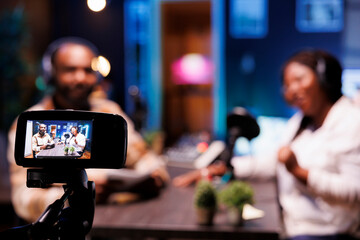 During live talk show broadcast on streaming platforms, cohosts use a professional camera for recording. African vloggers seen in blurry background using modern recording equipment to make a vlog.