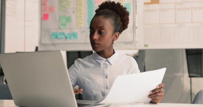 Focused African business woman working on computer, making research, comparing data, doing paperwork, working with papers, preparing financial report or legal documents sit at workplace desk in office