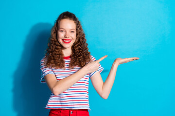Cheerful young woman pointing to copy space in striped shirt against blue background, showcasing...