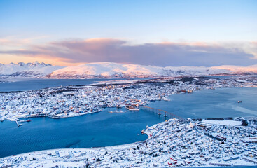 Aerial view of Tromso, Norway at sunset in winter from Storsteinen mountain. Tromsoya island with snowy roofs of Tromso downtown, harbor, Sandnessundet Bridge over Fjord and mountains in background