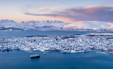 Aerial view of Tromso, Norway at sunset in winter from Storsteinen mountain. Tromsoya island with snowy roofs of Tromso downtown, harbor, cruise ship in fjord water and mountains in background