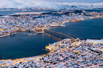 Aerial view of Tromso, Norway with city lights in winter from Storsteinen mountain. Tromsoya island with snowy roofs of Tromso town, harbor, Sandnessundet bridge over Fjord and mountains