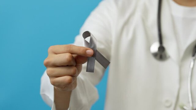 Young male doctor shows a gray ribbon, symbol of the fight against brain cancer, Parkinson's disease, asthma, blue background