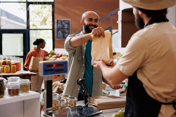 Smiling customer choosing organic products to buy and approaching vendor at counter to pay for bag...