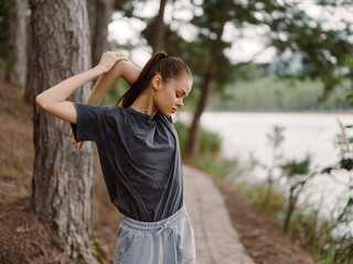 Young woman stretching outdoors by the lakeside, wearing a casual grey t shirt and sweatpants, enjoying nature and personal wellness Fitness and health concept