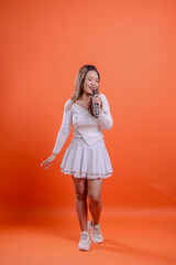 full length beautiful female white top standing singing cheerful close eyes candid hand holding audio mic while hand straight down isolated orange background. for fashion, advertising, entertainment