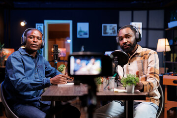 Portrait of vloggers seated at the table, recording video content using professional camera at home. Online talk show host having friendly interview with influencer, filming with device on tripod.