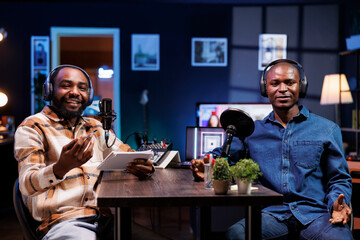 Two black men host a live talk show, discussing various topics while interacting with fans. They use professional microphones to create high-quality content for an online streaming service.