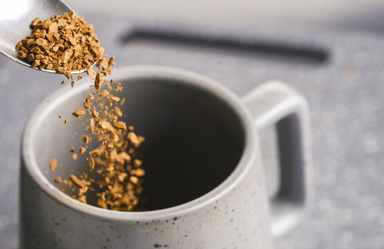 Grains of freeze dried instant coffee,falling from a silver tea spoon,into an empty cup,on a slate grey background..