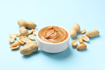 Tasty peanut butter in bowl and groundnuts on light blue background