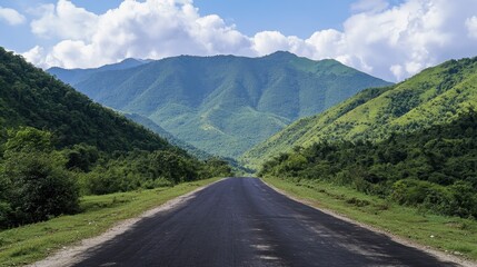 Naklejka premium A serene asphalt road surrounded by lush green mountains, under a clear sky with soft, white clouds on a sunny day.