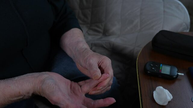 Retired person citizen measuring blood glucose levels by pricking fingertip in living room. Portable healthcare devices enabling self-monitoring for aging population.
