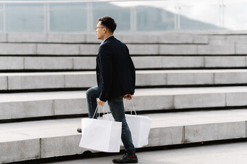 Man Carrying Shopping Bags Up Stairs: A young man confidently strides up concrete stairs,  carrying multiple white shopping bags in his hands. He is dressed in stylish, modern attire.