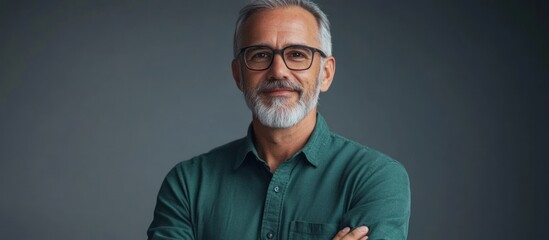 Fototapeta premium Portrait of a confident middle-aged man with a beard and glasses, standing against a gray background, with his arms crossed and a smile on his face.