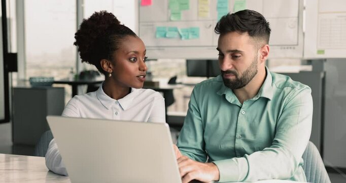 Two multinational teammates working on joint task, developing marketing strategy, share opinion and insights, look at laptop screen and learn corporate app, engaged in effective and friendly teamwork
