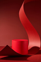 On a red backdrop, there is a lone red cylindrical podium. To create a dramatic image, a long strip of red paper is curled and stretched over the podium. A product presentation template picture.