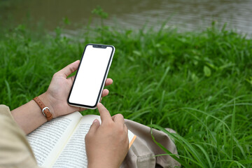Young woman using mobile phone while reading a book on green grass near serene lake