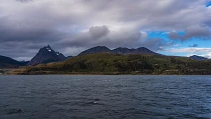 A picturesque mountain range. The Andes. Snow-capped peaks against a blue sky and clouds. Green vegetation on the coastal hills. The view from the ocean. The Beagle Channel. Argentina.Tierra del Fuego
