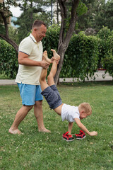 Happy father playing with his son in the park on a summer day. The dad is holding his child's legs...