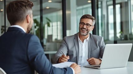 A professional and confident manager is having an interview with another man in the office, sitting at his desk while holding documents. 