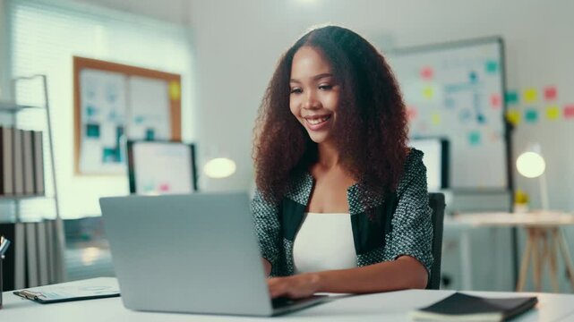 Young manager is sitting at her desk, smiling as she types on her laptop. She is surrounded by office supplies and appears to be in a good mood