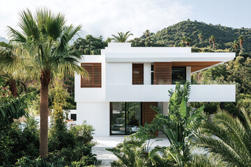Fototapeta premium A white modernist house with wooden details, surrounded by vegetation, plants, and palm trees, set against a backdrop of mountains in Andalusia. The photograph captures the architecture on a sunny day