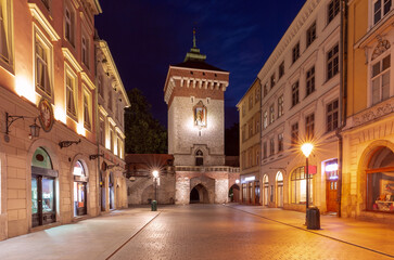 Fototapeta premium Florianska Gate at night in Krakow, Poland. The illuminated medieval tower is surrounded by historic buildings along Florianska Street.