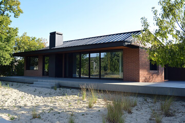 A small, single-story house with an exterior of red brick and a black metal roof stands on the sandy ground, surrounded by trees. The walls feature large windows that blend into the nature outside.