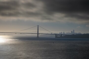 Dark low clouds and bright sun over suspension bridge with city in distance