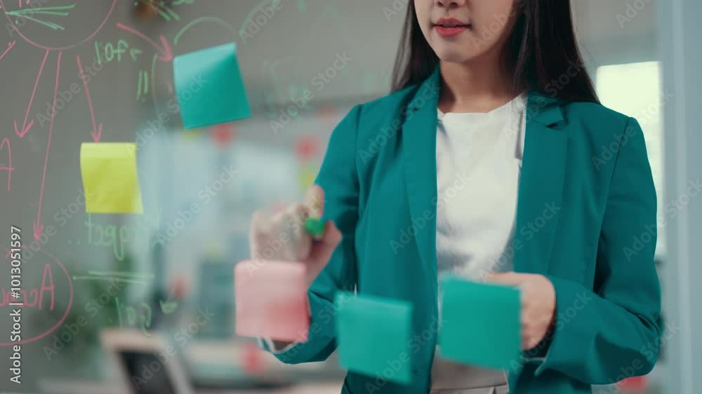 Poster businesswoman in a teal blazer writes on sticky notes while organizing thoughts on a glass wall, sho