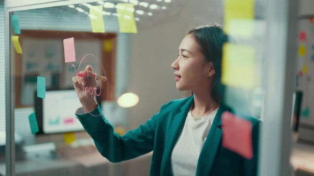 Businesswoman in a green blazer draws a flowchart on a glass wall, actively engaging in a brainstorming session in a modern office environment. Creativity and strategy come together