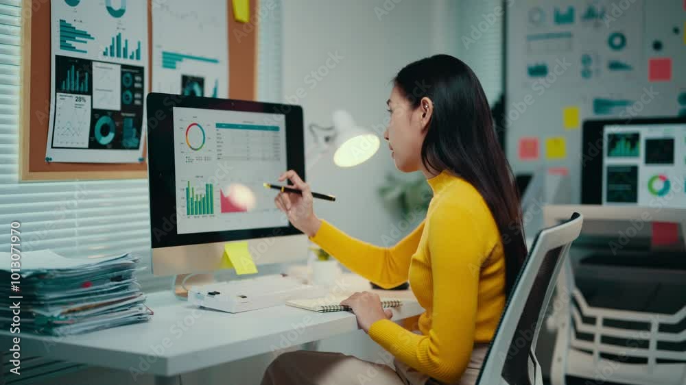 Poster young asian businesswoman is analyzing financial data on a computer screen in her office. she is tak