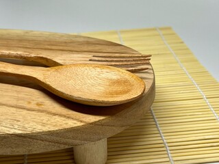 A wooden spoon and fork placed on a round wooden board against a bamboo mat background.