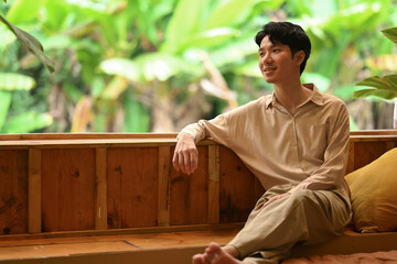 A man lounges peacefully indoors, barefoot, with a lush green background. His relaxed posture invites a sense of calm and contentment.