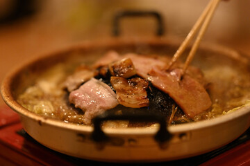 Close-up of Slices of pork cook on the hot Moo Kata grill, releasing a savory aroma as the meat sizzles and the flavorful broth below bubbles with vegetables and seasonings