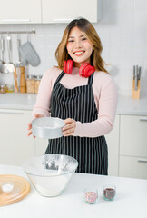 Young happy Asian beautiful woman, female wearing aprons intends and headset standing in kitchen sifting flour for homemade bakery, bread baking making pastry or cake. Lifestyle enjoy cooking at home