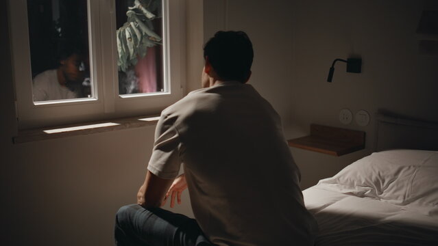 Sorrowful man sitting troubled at night bedroom reflected in dark window closeup