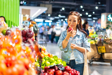 Beautiful Asian mature woman choosing and buy fresh fruit and vegetable with using reusable mesh cotton shopping bag at shopping mall supermarket. Sustainable living and healthy lifestyle concept.
