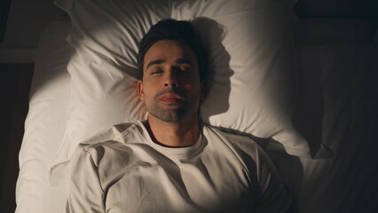 Fototapeta premium Guy lying dark bedroom at white bedding looking up closeup top view. Man on bed