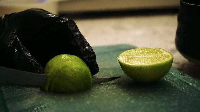 Slicing a green lime with a sharp knife. Lime wedges on a green cutting board. Cooking process, part of a hand in black gloves.