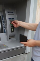 A man is using a card in the ATM machine while holding his wallet with the other hand.