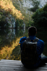 A young guy with backpack sits on a wooden pier on the shore of a mountain lake and enjoys the silence, tranquility and views of nature alone. Reflection of the autumn forest and rocks in the water