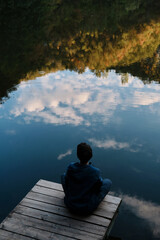 A young woman with short haircut sits on a wooden pier near mountain lake and enjoys the silence, tranquility and views of nature alone. Reflection of the autumn forest and sky in the water.