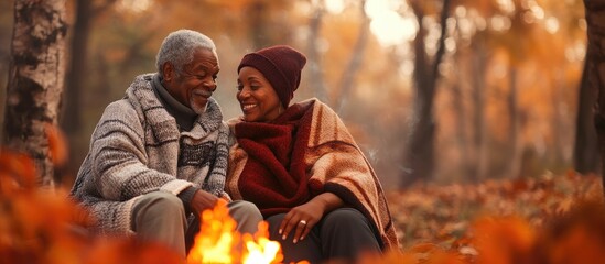 camping and hiking tourism, autumn vacation, active lifestyle. senior african american couple next to campfire in forest, surrounded by red leaves, covered with cozy blanket. banner with copyspace