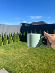 A person holds a coffee mug in a sunny backyard with green grass, trees, and a clear blue sky in the background. Peaceful morning outdoors.