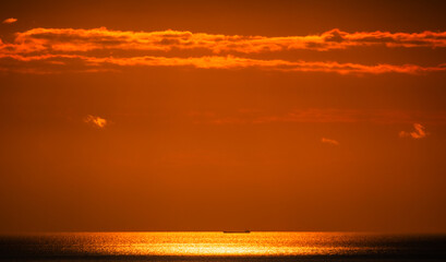 A breathtaking sunset casts a warm golden hue across the tranquil ocean, while a lone cargo ship sails peacefully on the horizon, creating a serene atmosphere.
